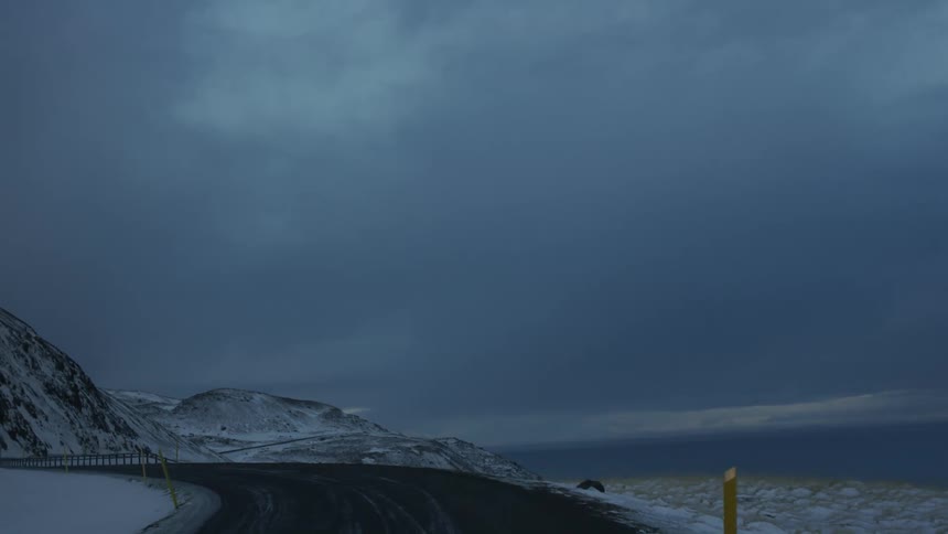 A winding mountain pass curves around a snowladen slope with a guardrail visible on the roads edge under a darkening sky