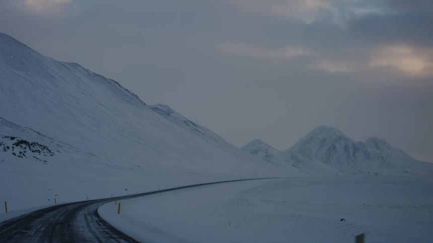 A winding mountain road curves through a snowy landscape flanked by bright yellow markers beneath a subdued winter sky