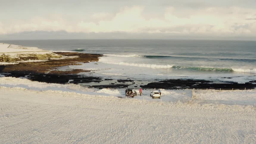 Aerial shot of some people around two cars in a snow covered area on a beach