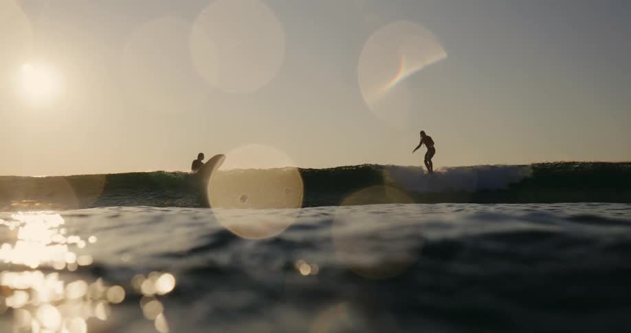 Two surfers riding a gentle wave at sunset with the warm golden light casting a dreamy glow and lens flares adding to the serene ocean atmosphere