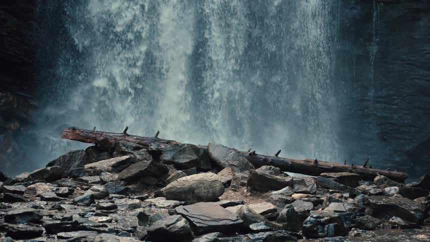 Water falls down in front of a rock wall and disappears behind a fallen tree trunk on a pile of large flat rocks