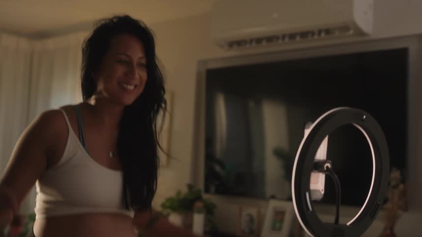 A pregnant woman in a white tank top is dancing energetically recording or streaming an exercise session from a cozy living room