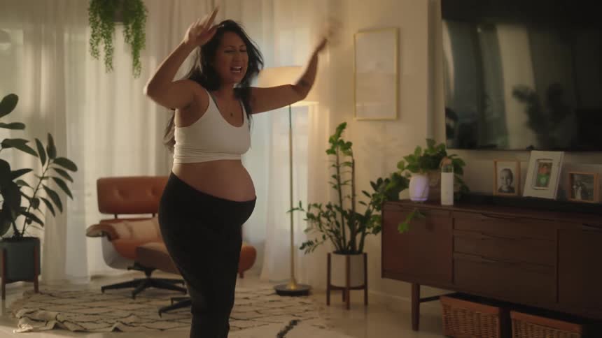A pregnant woman in a white tank top is dancing energetically recording or streaming an exercise session from a cozy living room