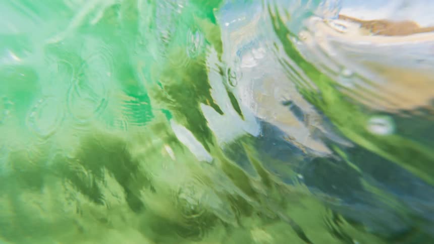 From an underwater perspective a waves turbulent surface is visible churning with white foam and lighter shades against a darker ocean floor