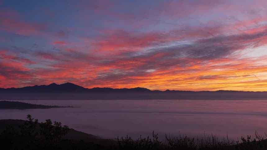 A timelapse from a mountaintop vantage reveals rolling clouds and fog filling the valley below against a vibrant sky