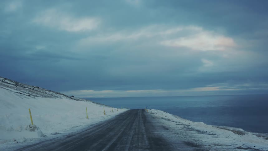 A mountain road stretches straight ahead beside a steep snowy slope with street poles on the left and the ocean beyond