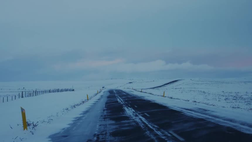 A desolate snow covered highway stretches into the distance under an overcast sky the dark asphalt contrasting sharply with the white landscape and a fence line running parallel to the road