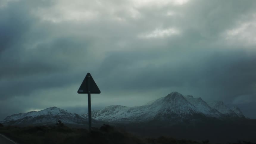 A road sign stands in the foreground against a backdrop of snow capped mountains and a cloudy overcast sky