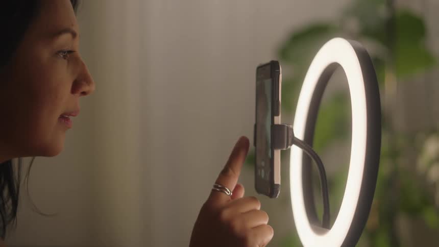 A woman adjusts a smartphone mounted on a ring light presumably for recording or video conferencing