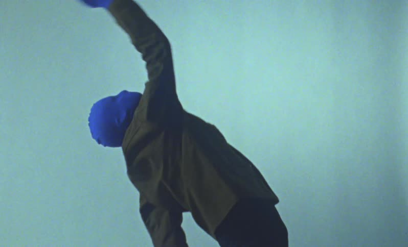Person with a blue hood covering their entire face and wearing a jacket dancing in a studio