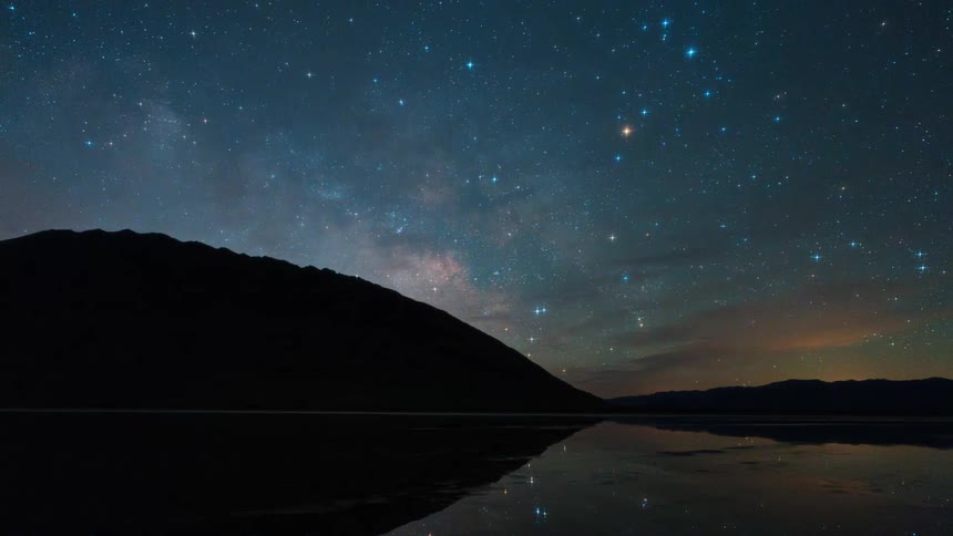 A silhouetted mountain range is reflected in the still water of a lake under a vast twinkling night sky filled with countless stars