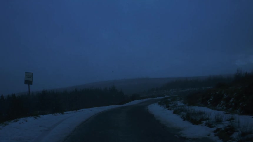 A dark snowy road is seen through a car windshield with a wiper blade partially obscuring the view and a road sign is visible in the distance