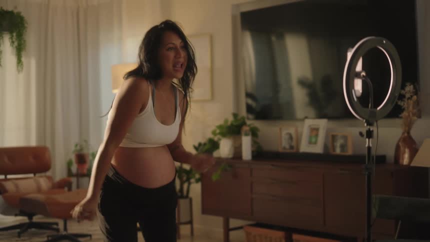 A pregnant woman enthusiastically dances in front of a ring light and smartphone for a video recording in her living room