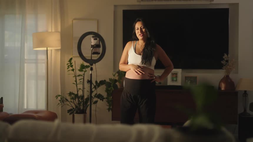 Pregnant woman wearing a light top and black skirt is in a bright room interacting with a cell phone with a ring light in the background