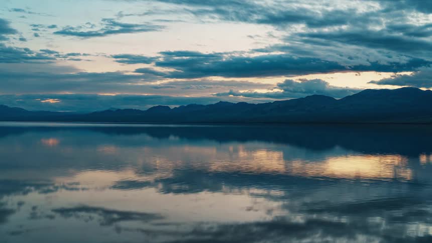 A tranquil lake mirrors the softly lit clouds and silhouetted mountains in a serene twilight setting