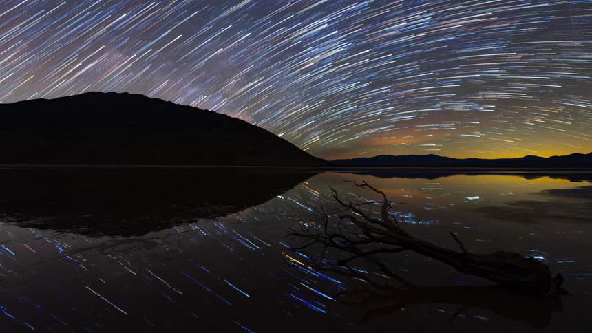 Star trails curved across the night sky over a calm lake reflecting lights in the sky with a dark mountain and a submerged branch in the foreground