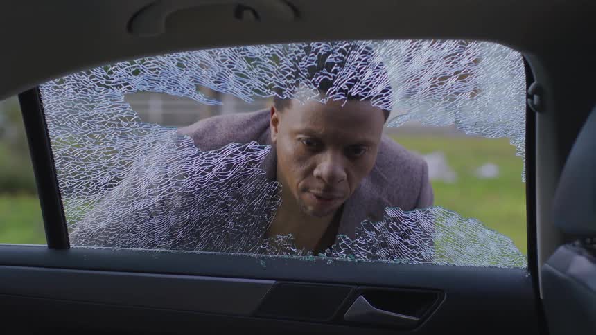 Man examines a broken car window and the baseball responsible on a summer day