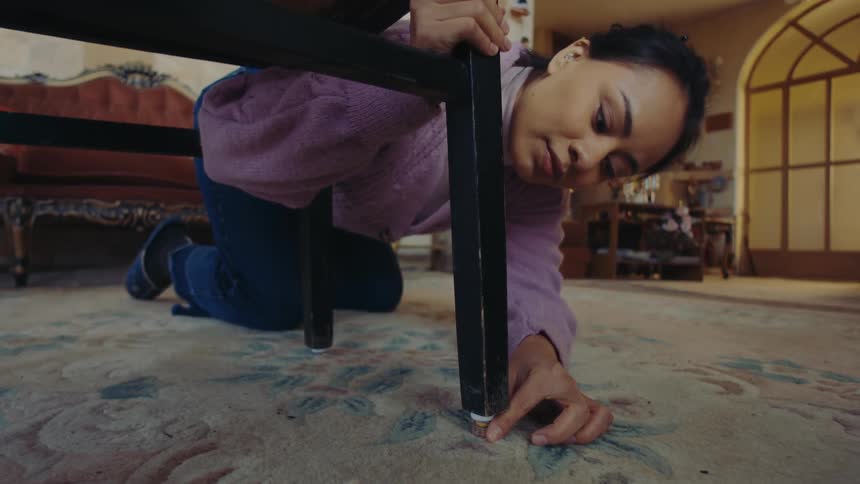 A woman kneeling on a patterned carpet reaches under a chair to place a piece to raise the leg and balance the chair