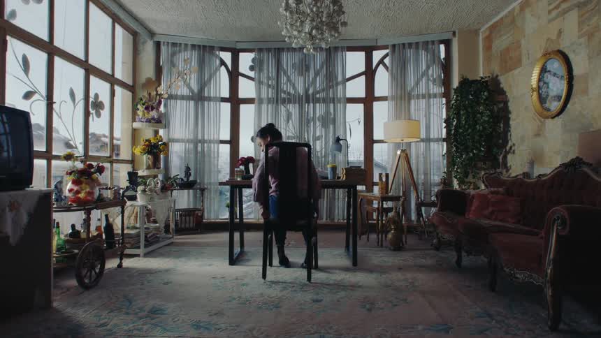 An asian woman in a violet wool sweater and blue jeans enters a home office with big windows overlooking the city sits on a chair stands to adjust it and then sits back down with her hair in a bun