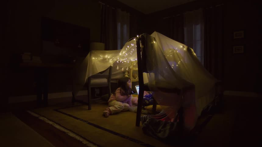 In a dimly lit room two children are inside a makeshift fort decorated with string lights