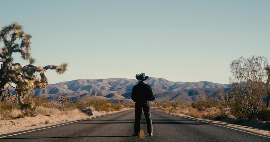 Man stands in the center of a desert highway leading towards the mountains