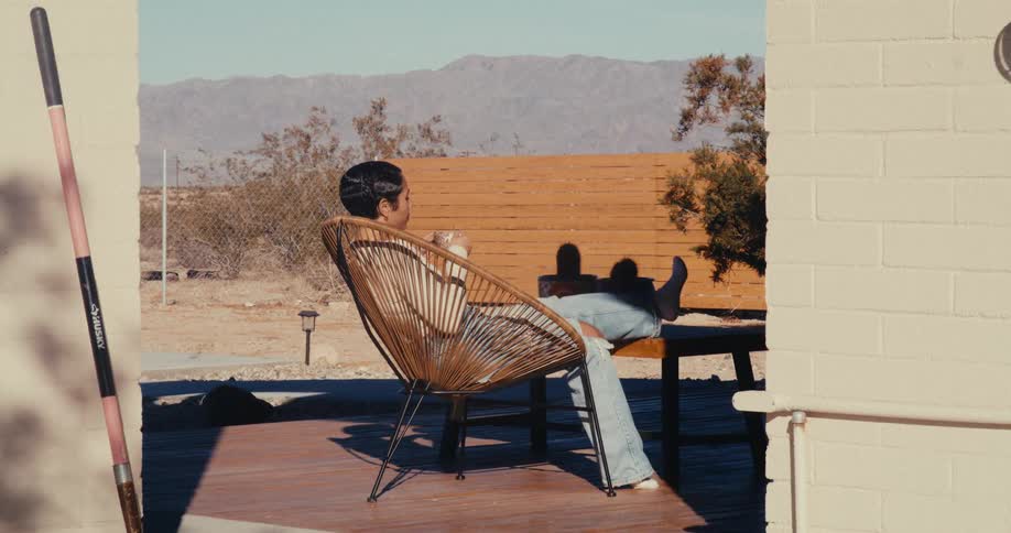 A woman is sitting on a wooden deck in a wicker chair drinking from a cup with a desert landscape and mountains in the background