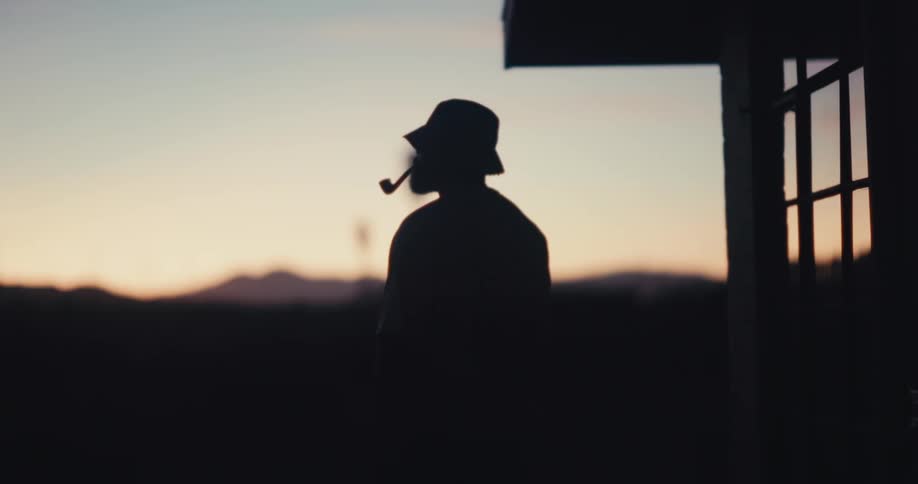 Man smokes a pipe while walking back and forth against a red tinted horizon in the dark