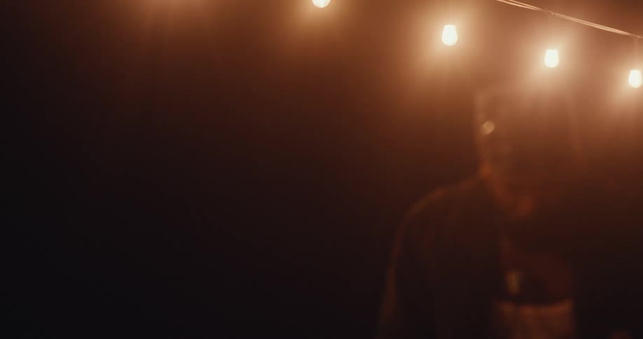 A person wearing a bandana is dancing outdoors at night under a string of illuminated light bulbs