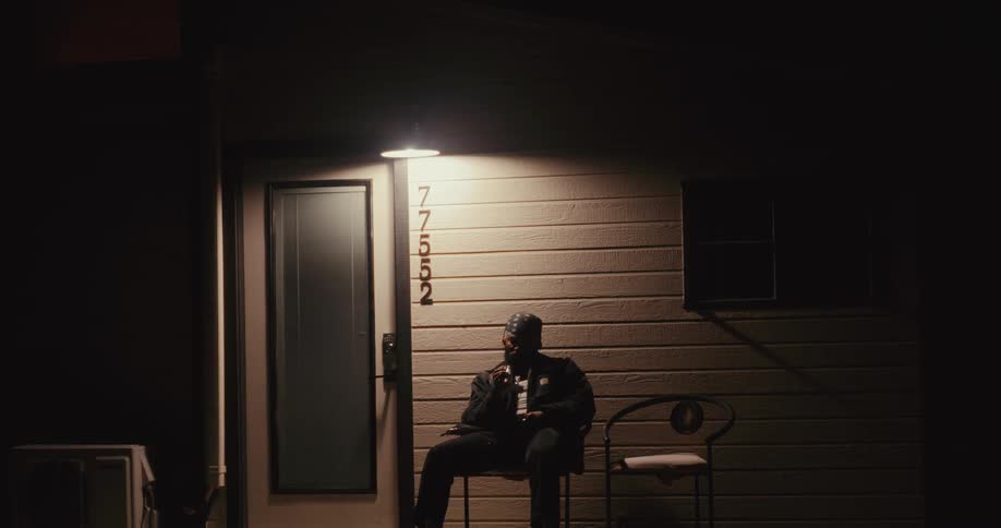 A man is leaving a wooden house and sitting on a chair on the porch and then gets up and walks forward and it is night