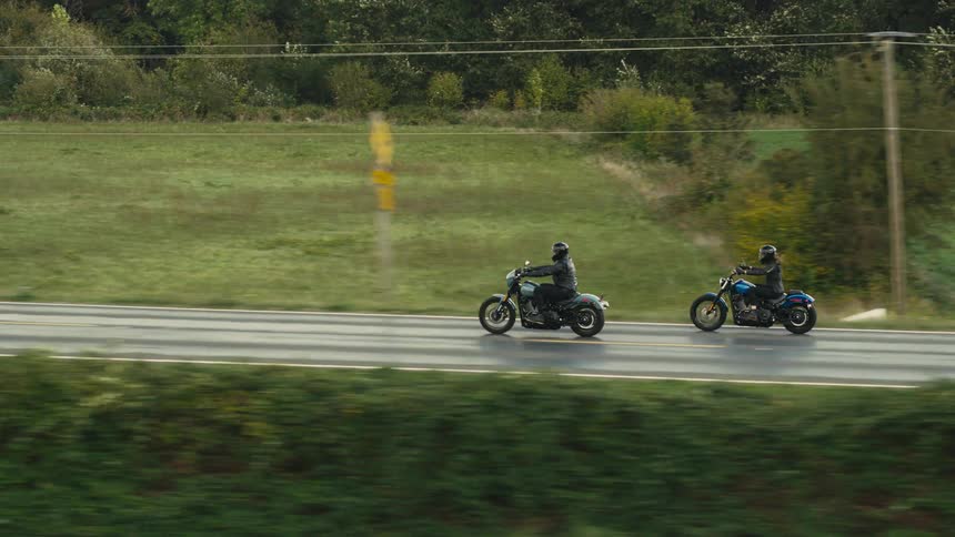Two motorcyclists ride on a road surrounded by green fields and trees