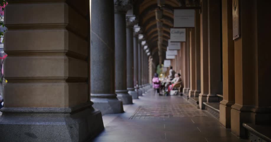 A long covered walkway with a series of columns on both sides leads to a distant group of people sitting at a table