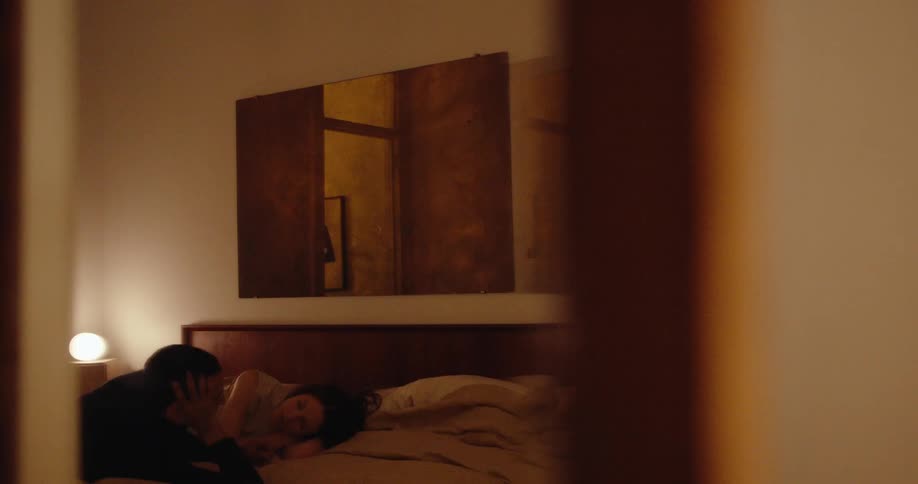 A dimly lit bedroom scene shows two people lying on a bed with one person resting their head on the others shoulder