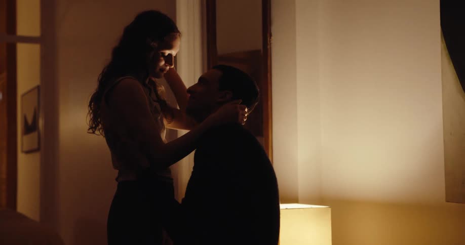 A woman standing hugging a man who is sitting on a bed in an interior with a lamplight