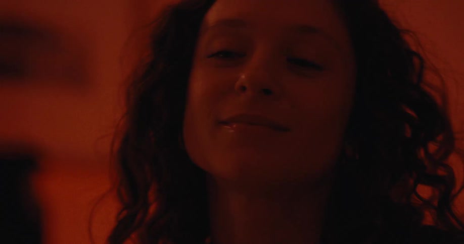 Man and woman interact in a room lit by red lighting before she starts laughing