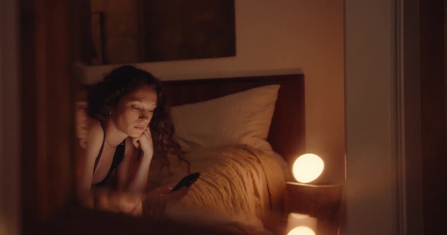 A woman is laying her back on the bed scrolling on her phone and then rolls over to her stomach