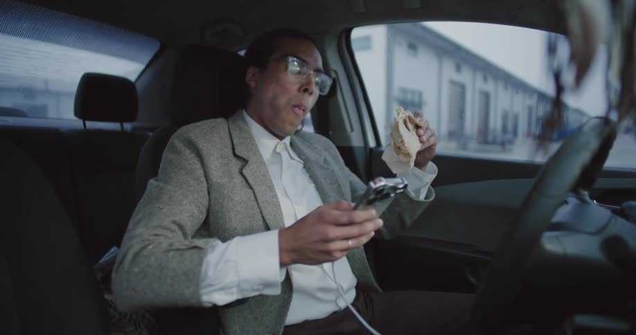 A person is sitting in the drivers seat of a car wearing a gray suit jacket and white shirt holding a sandwich in one hand and a smartphone in the other