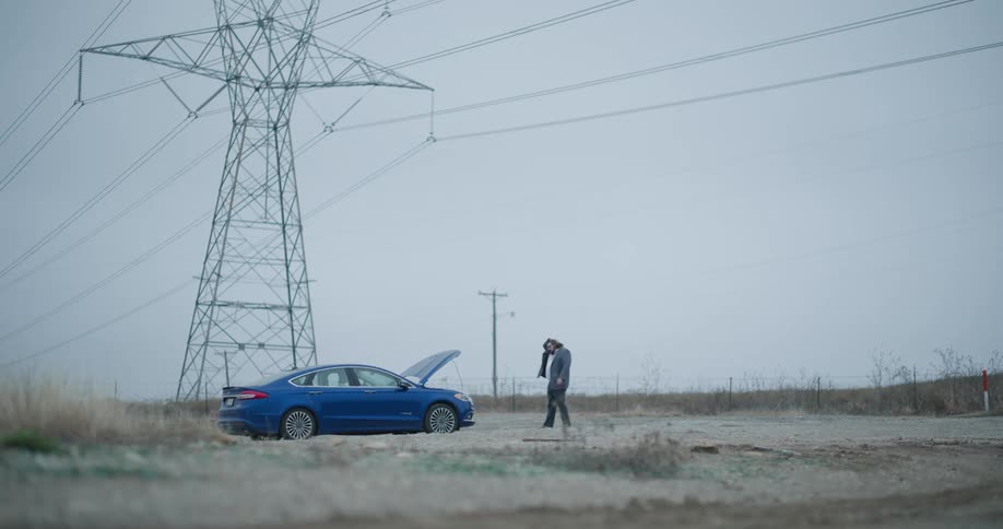 Man kicking a blue car with the hood open on a dirt road near an electrical transmission tower