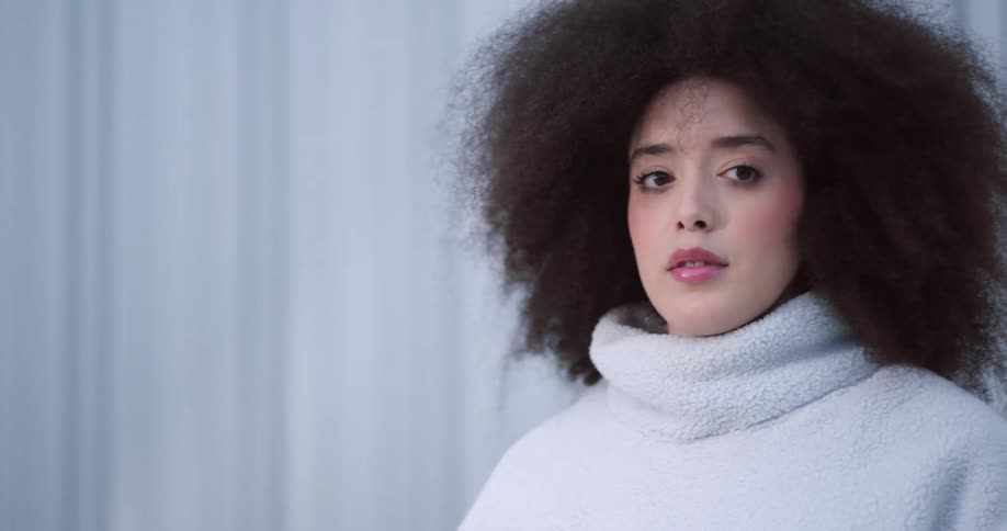 A young woman with voluminous curly hair is wearing a white fuzzy sweater and talking on her phone with a calm expression standing in front of a metallic background