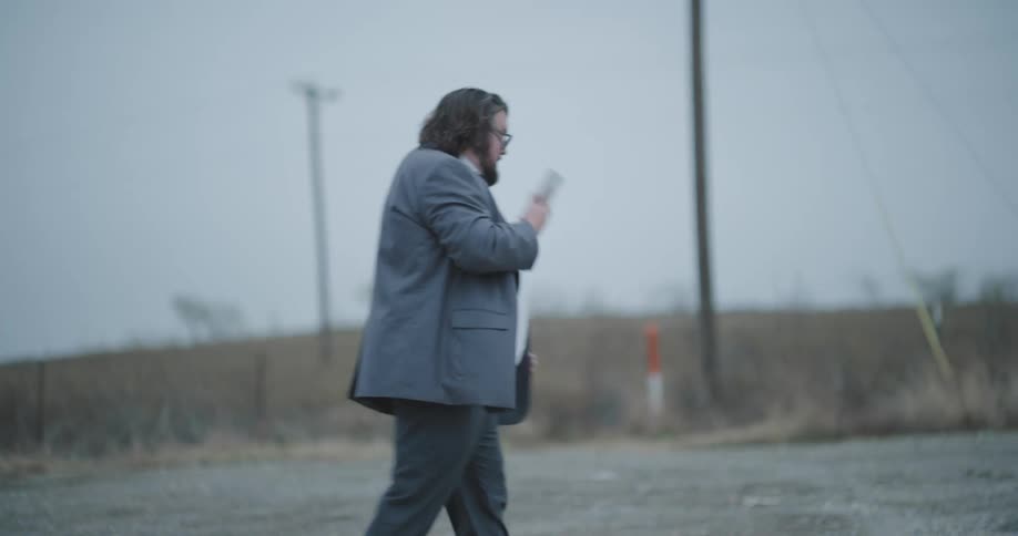 A man in a suit with disheveled hair and glasses talks on his phone while leaning over the open hood of a car on the side of a rural road he gets mad and hangs up in frustration