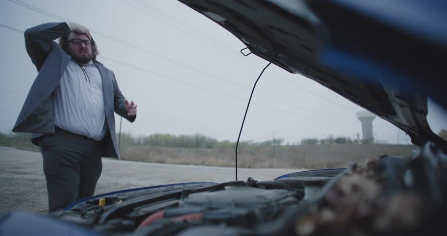 A man in a suit with disheveled hair and glasses leans over the open hood of his broken down car on the side of a rural road consumed by rage