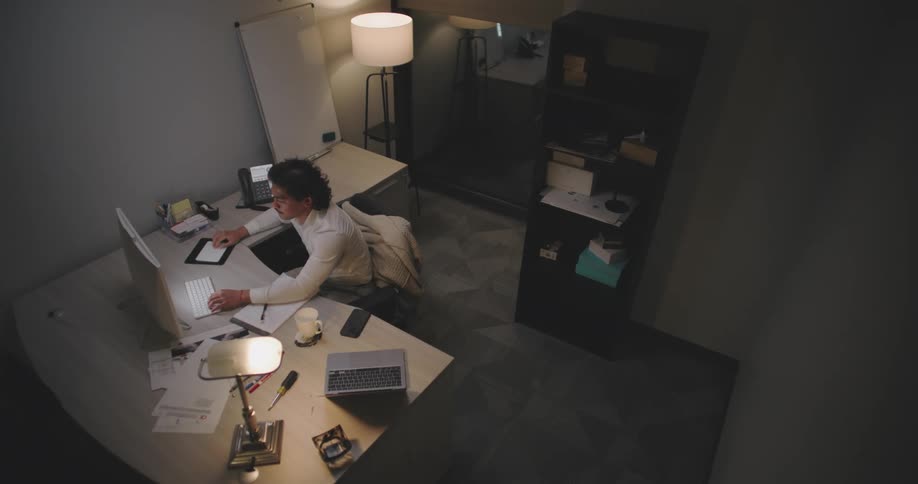 A man is sitting in a dimly lit office leaning back in his chair while surrounded by a cluttered desk with papers a laptop a tablet and a cup of coffee he works on papers then crumples them in frustration and tosses them away
