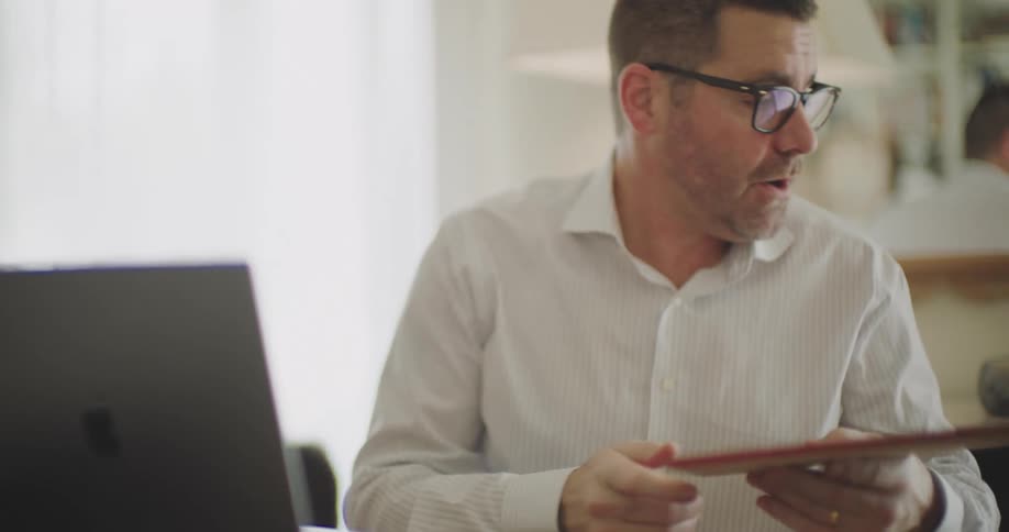 A father in a white shirt works from home on a laptop while interacting with his children in a brightly lit room