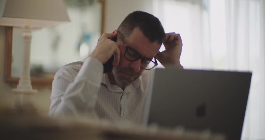 A person in a white shirt sits at home working on a laptop while making a call on a smartphone