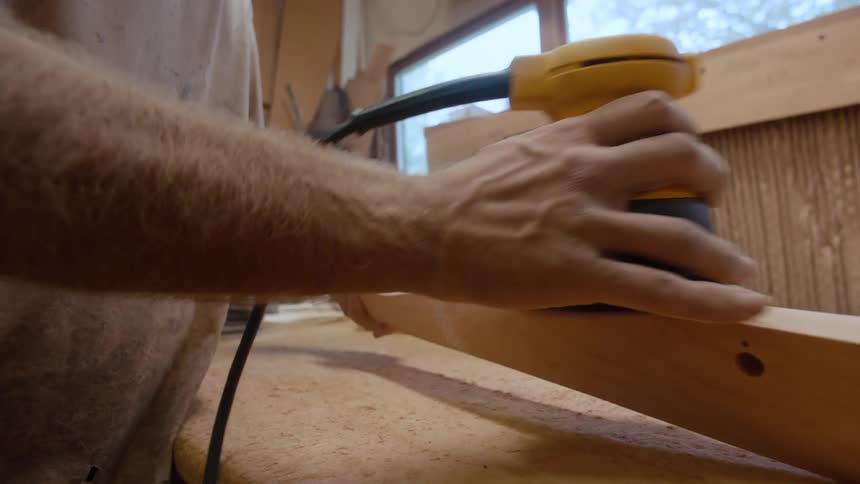A whitebearded man wearing a mask and using a tool to scrape the sides of a wooden slat
