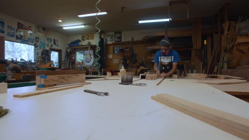 A man in a blue shirt and wearing a mask is passing a wooden slat through a machine that makes the cut