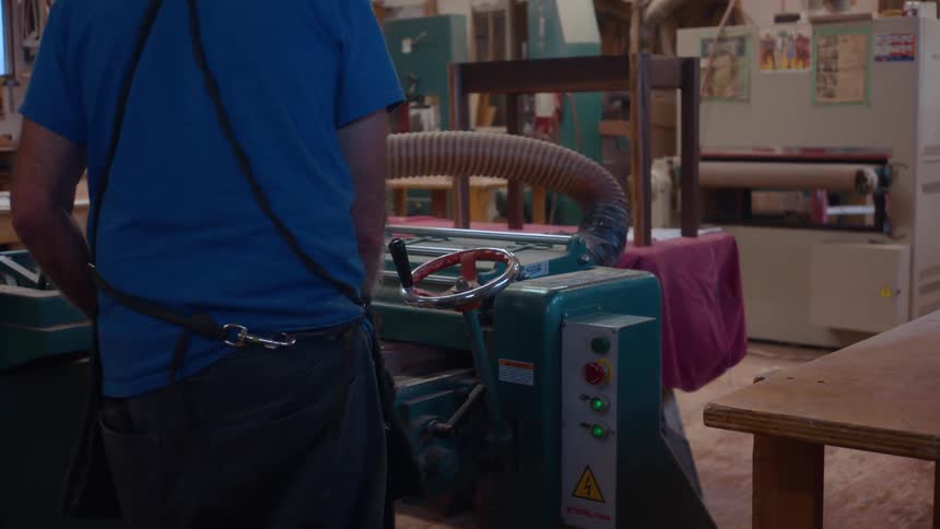 A person is operating a green industrial woodworking machine with a large hose attached in a workshop filled with various tools and equipment