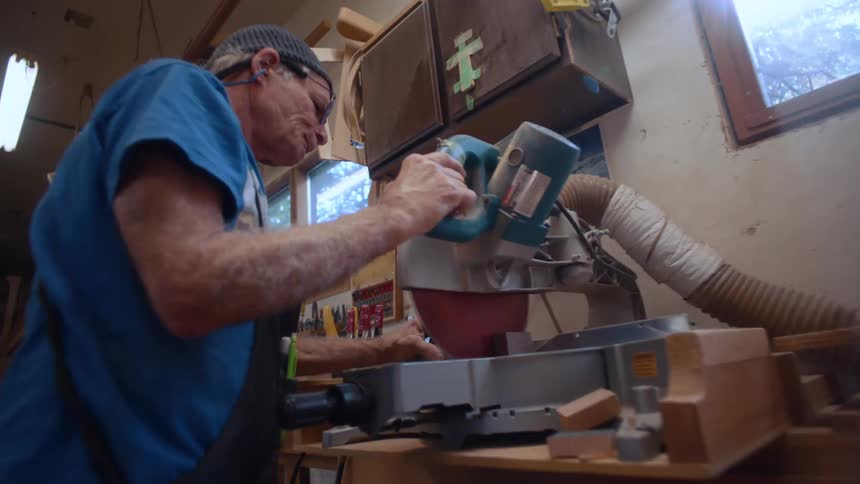 A person is operating a miter saw in a woodworking shop filled with various pieces of wood and tools