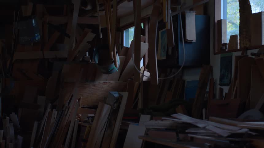 A dimly lit woodworking shop is filled with scattered planks and frames creating a cluttered scene