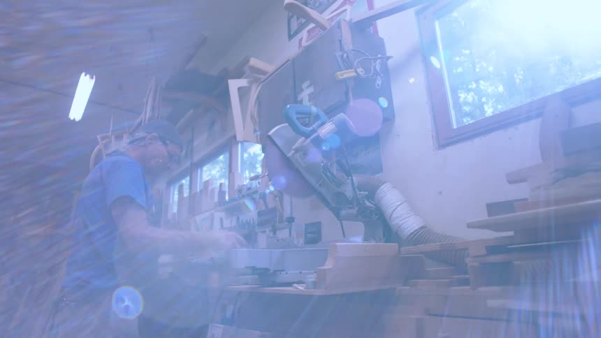 A person is operating a miter saw in a woodworking shop filled with various pieces of wood and tools