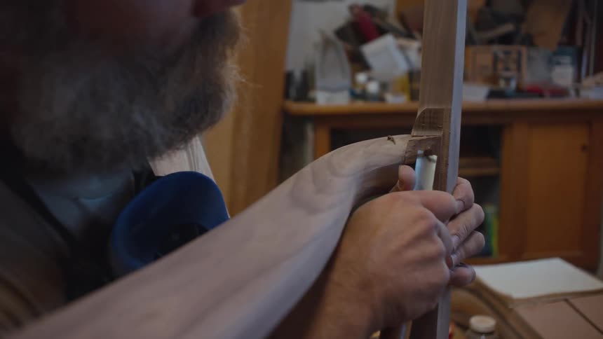 A person with a beard is carefully working on a wooden object in a workshop using both hands to hold and adjust a piece of wood